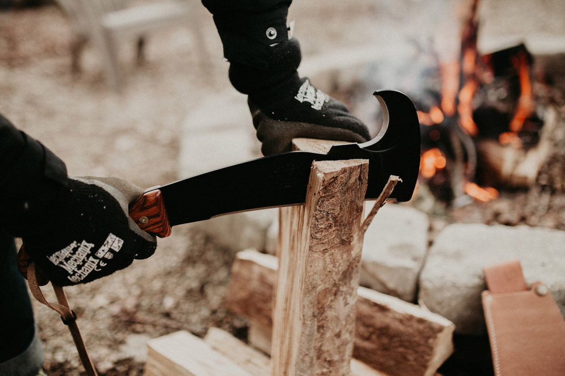 Person wearing work gloves using a Woodman’s Pal machete to split firewood near a campfire outdoors.
