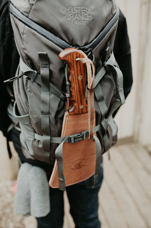 A close-up of a gray bug-out bag/backpack with a Woodman's Pal survival tool secured in a leather sheath attached to the exterior straps.