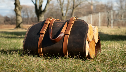 A Woodman’s Pal leather log carrier bag filled with firewood, resting on grass in an outdoor setting with trees in the background.