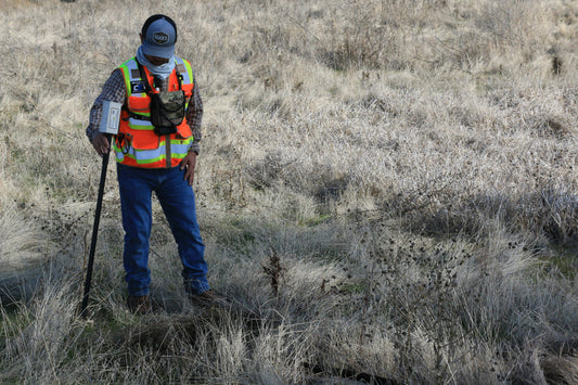 A surveyor placing equipment in a field covered in overgrown weeds and brush.