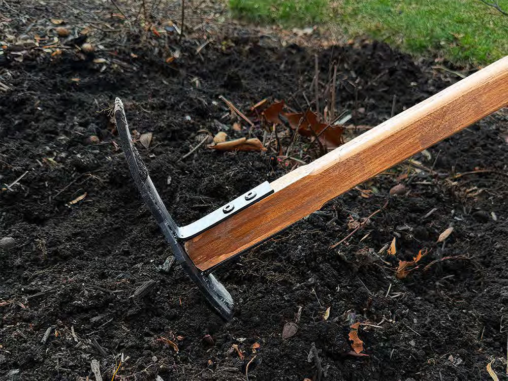 Woodman’s Pal heavy-duty gardening hoe cutting into dark, compacted soil in a garden bed, showing how the tool helps break up dense ground.