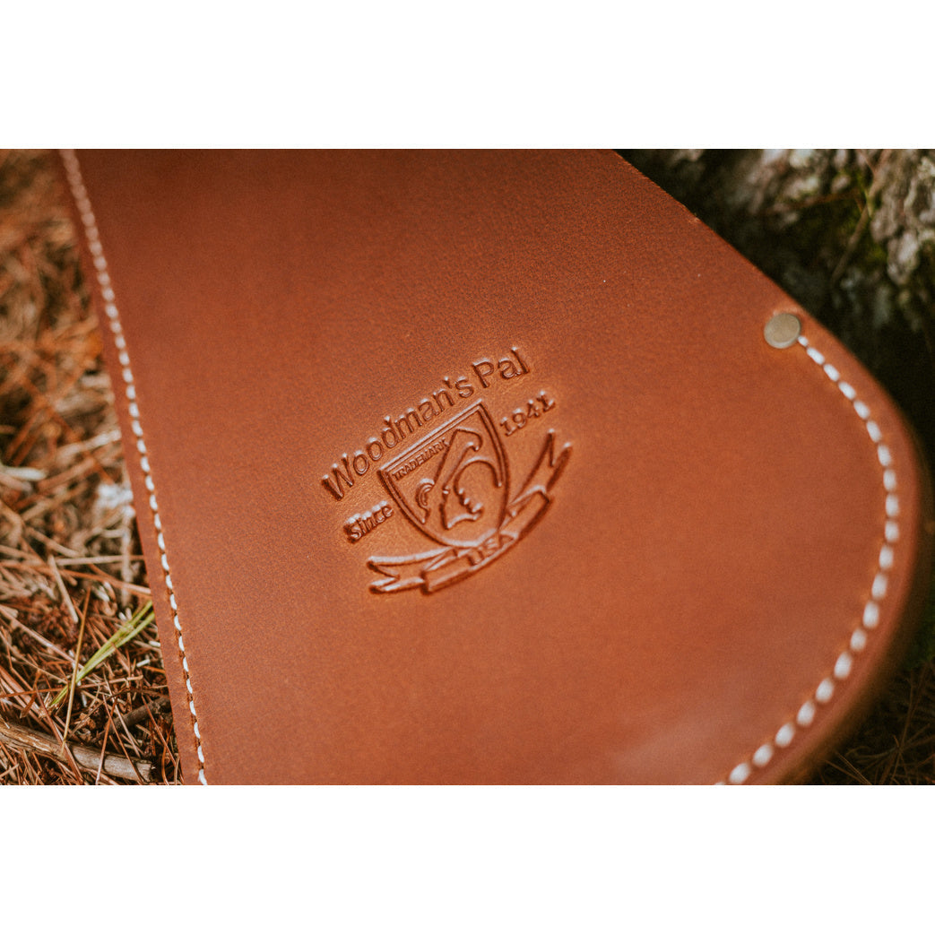 Close-up of a brown Woodman’s Pal leather machete sheath with white stitching and metal rivets resting on pine needles beside a tree.