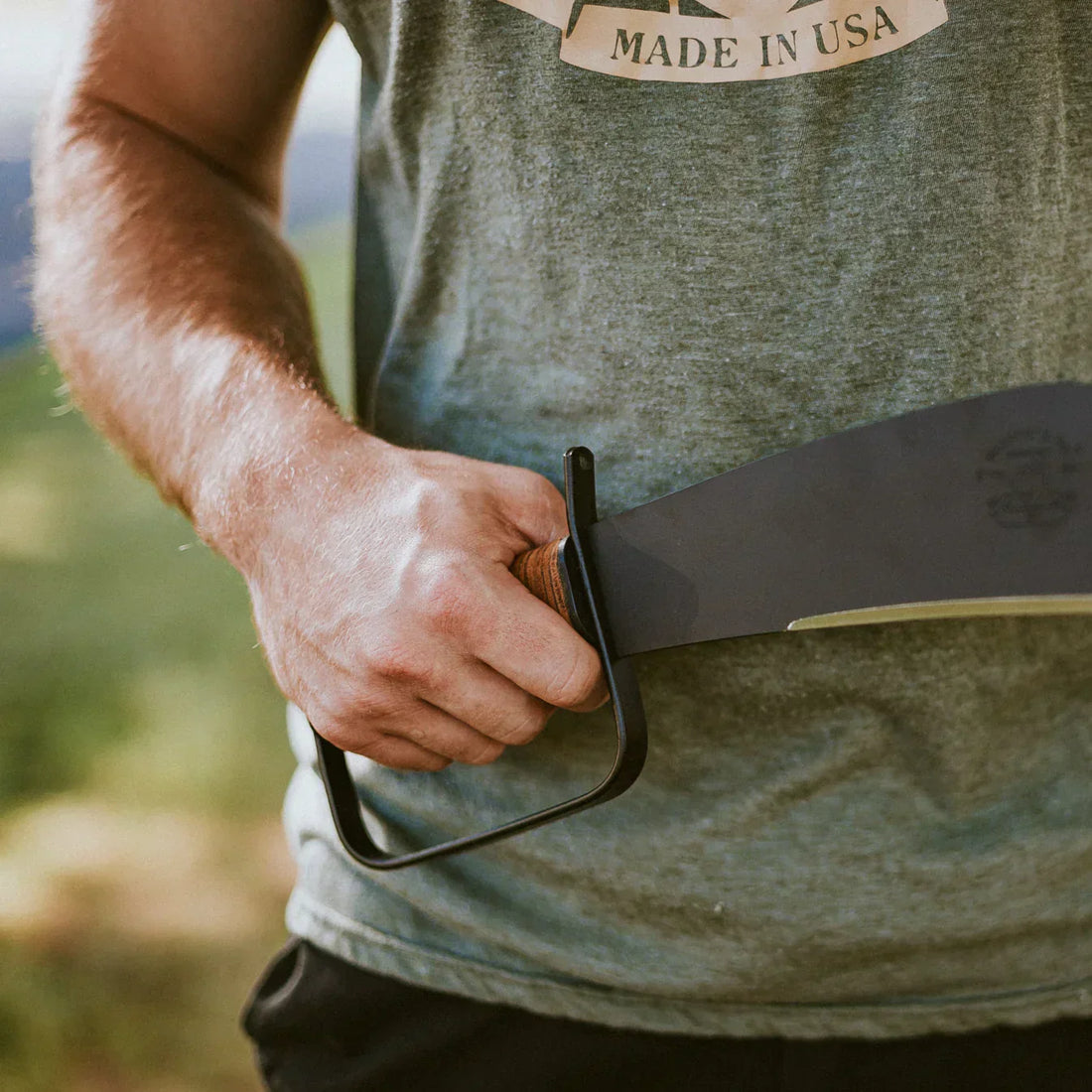 Person holding a Woodman’s Pal machete with a leather-wrapped handle and steel hand guard outdoors.
