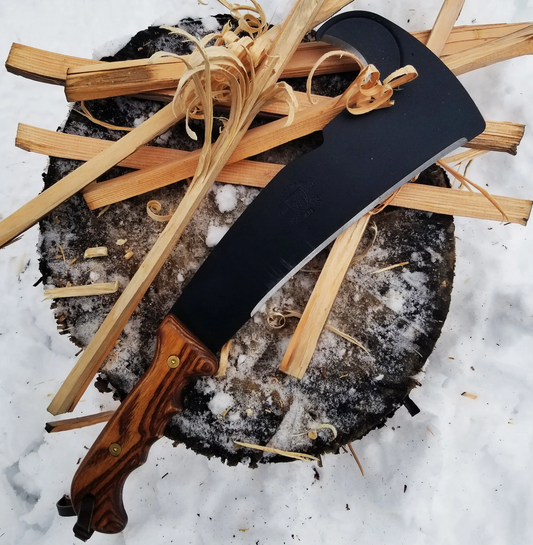 A Woodman’s Pal machete with a wooden handle rests on a snowy tree stump surrounded by freshly split kindling and wood shavings.