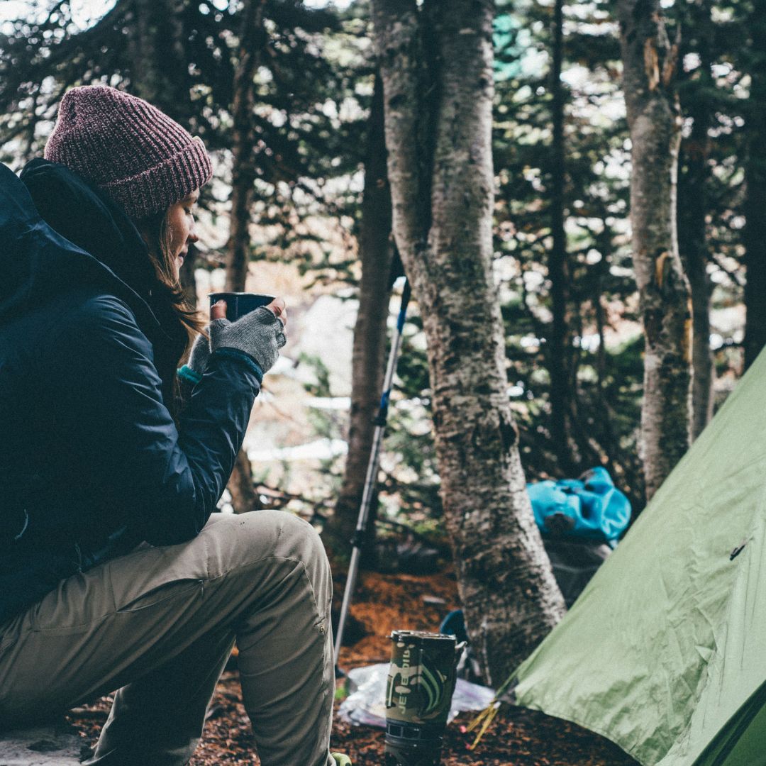 A camper dressed in cold weather gear, sitting next to a tent holding a camping mug filled with coffee. 