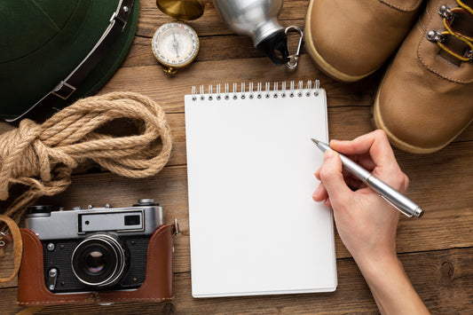 A person writes in a blank field journal surrounded by outdoor gear including a compass, rope, canteen, boots, a hat, and a camera on a wooden table.