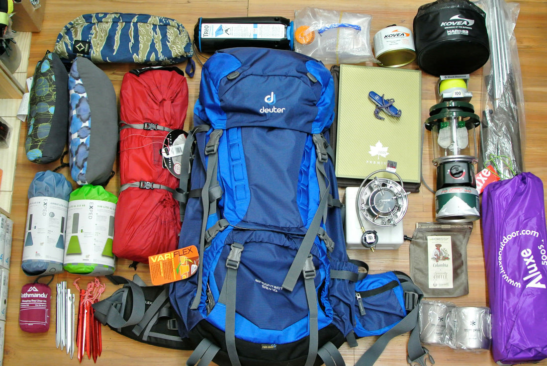 Flat lay of a packed hiking backpack surrounded by organized camping gear including a tent, sleeping pad, stove, cookware, fuel canister, lantern, stakes, and accessories on a wooden floor.