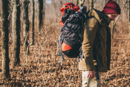 A camper wearing a red beanie and green jacket carrying a large backpack while hiking through a leaf-covered autumn forest.
